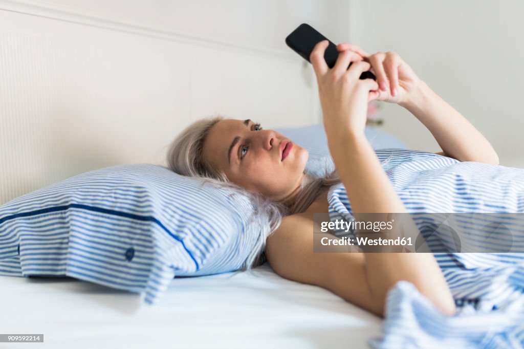 Serious young woman lying in bed checking cell phone