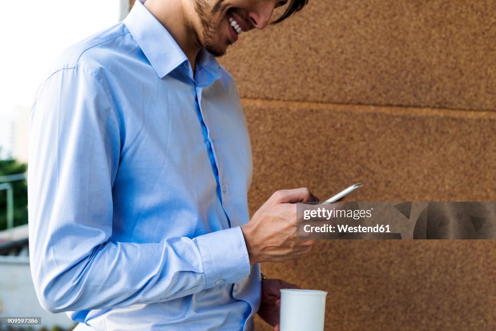 Laughing businessman standing on balcony with coffee mug using smartphone, partial view