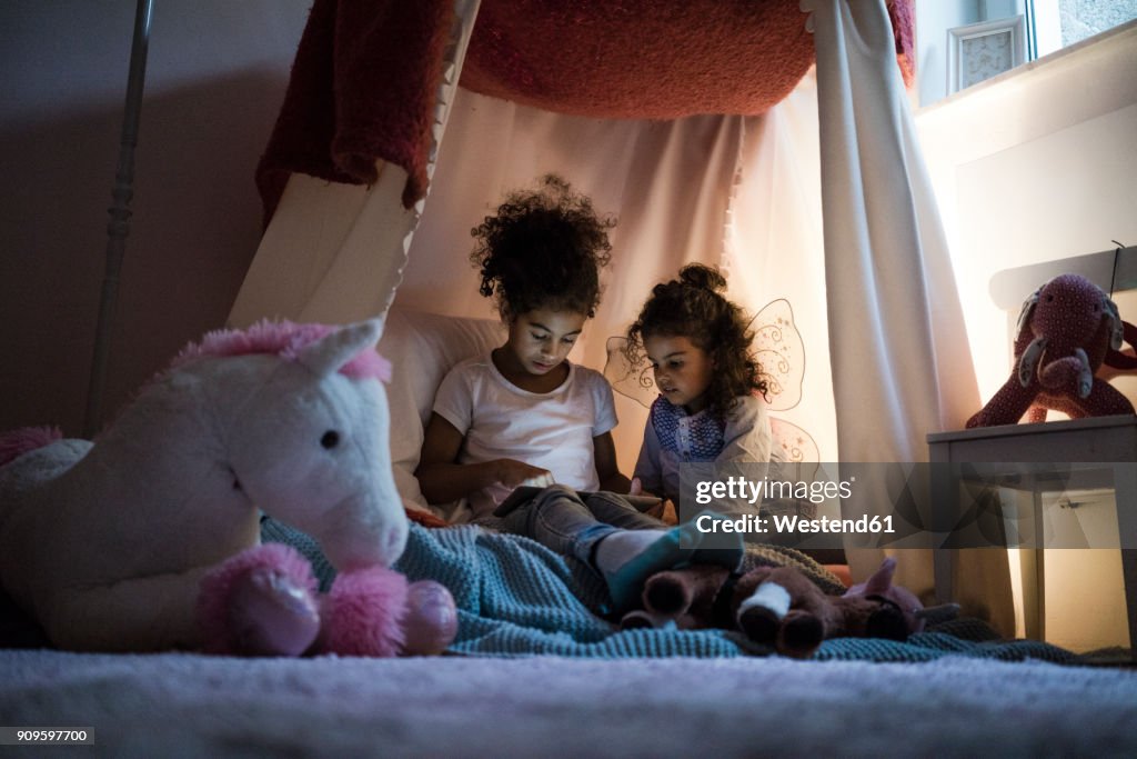 Two sisters sitting in dark children's room, looking at digital tablet