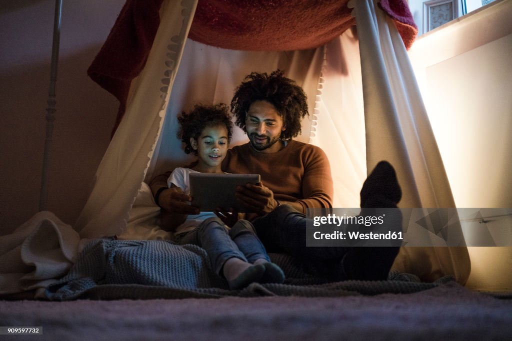 Father and daughter sitting in dark children's room, looking at digital tablet