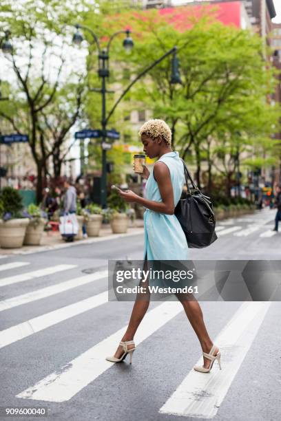usa, new york, young blonde african-american woman with cup of coffee and smart phone crossing street - businesswoman nyc stockfoto's en -beelden