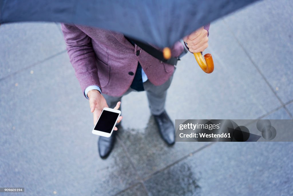 Businessman standing under umbrella holding cell phone, partial view