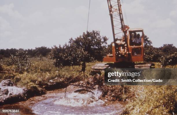 Photograph of a dragline cleaning debris from a water pool accumulated in a drainage canal amid vegetation, ensuring water flow and flood control as...