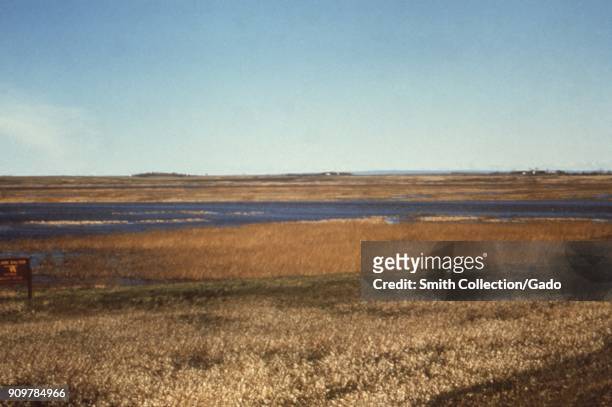 Photograph of a landscape consisting of a river with reed and grassland around, clear blue sky above and a sign, at J Clark Salyer National Wildlife...