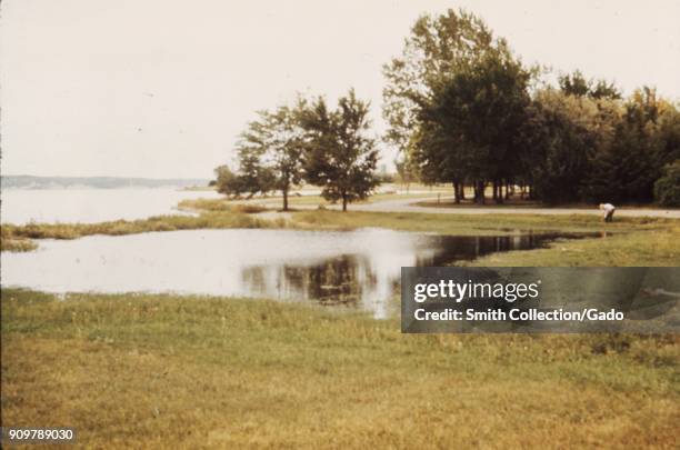 Landscape photograph of a lake reservoir, including an area of seepage in a flat, grassy area with trees, one person, pedestrian path and distant...