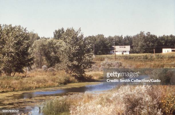 Landscape photograph of a U-shaped oxbow bend in a river, with trees, two white buildings and overhead power lines visible in the background, taken...