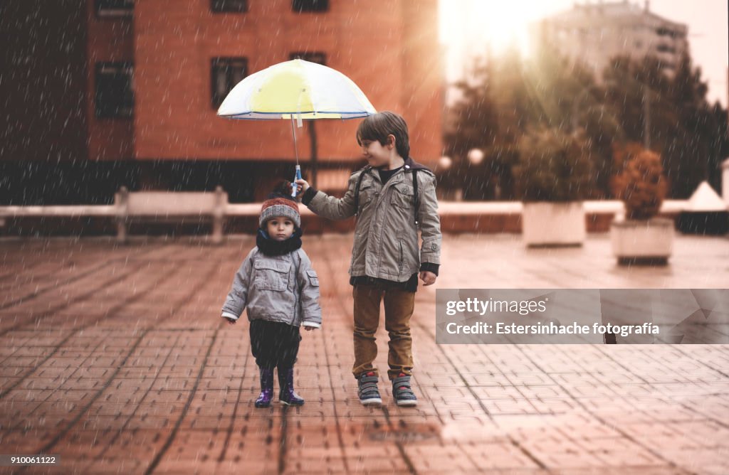 A little boy and his older brother protect themselves from the rain with an umbrella, in the city