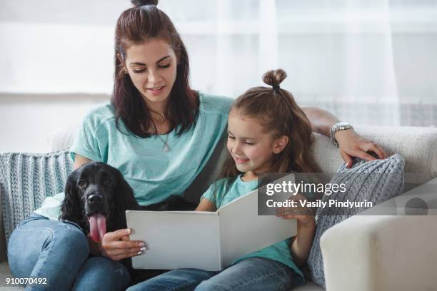 girl reading book by mother and dog on sofa at home - messy hair bun stock pictures, royalty-free photos & images