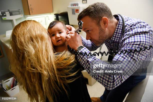 Physician's assistant Ryan Conrad, right, looks inside the ear canal of patient America Montes, 8-months-old, as her mother Nancy Espino, left, holds...