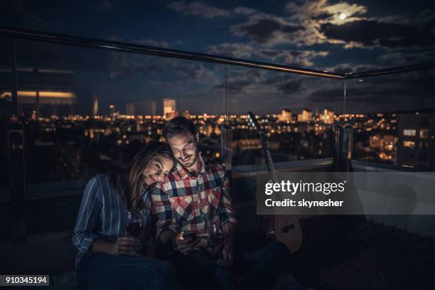 junge lächelnde paar entspannende auf ein penthouse-terrasse bei nacht und mit handy. - penthouse stock-fotos und bilder