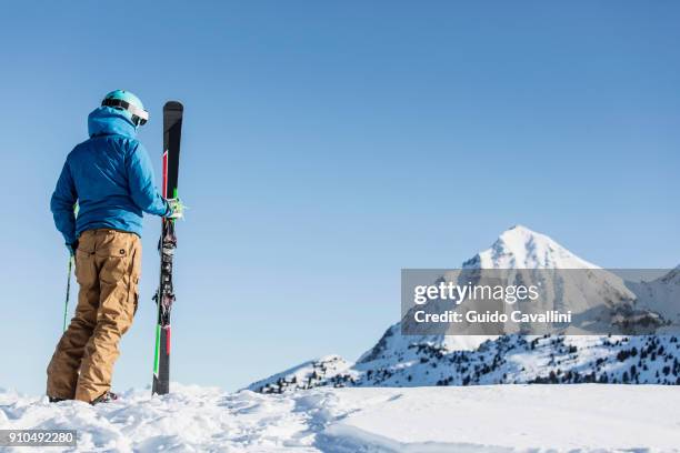 skier standing in snow, holding skis, looking at mountain, rear view - ski wear stock pictures, royalty-free photos & images