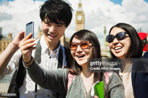 smiling man and two women with black hair taking selfie with smartphone, standing on westminster bridge over the river thames, london, with the houses of parliament and big ben in the background. - big ben selfie stock pictures, royalty-free photos & images