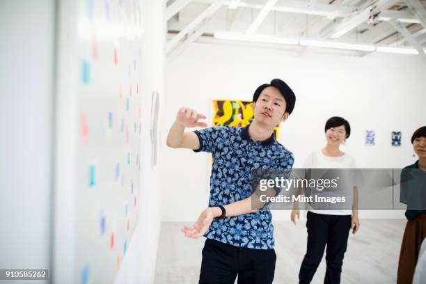 man wearing blue shirt standing next to modern painting on white wall in art gallery, two women standing in background.. - art and craft product stock pictures, royalty-free photos & images