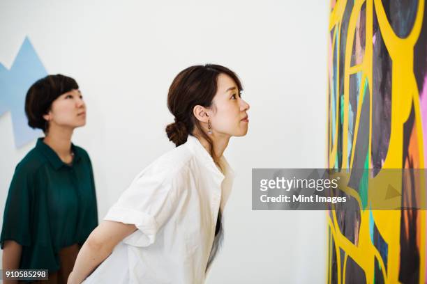 two women standing in an art gallery, looking at an abstract modern painting. - voyant photos et images de collection