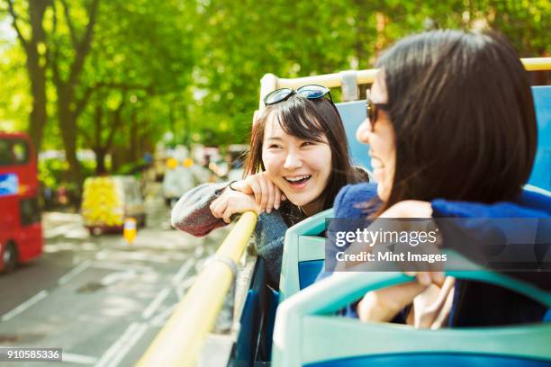 two smiling women with black hair sitting on the top of an open double-decker bus driving along tree-lined urban road. - tourist bus stock pictures, royalty-free photos & images