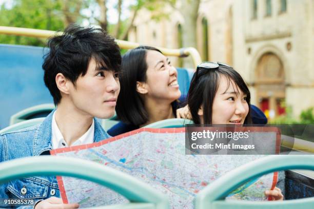 smiling man and two women with black hair holding city map, sitting on the top of an open double-decker bus, driving along urban road. - woman sitting top man stock pictures, royalty-free photos & images