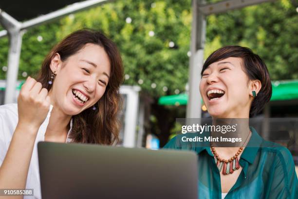 two women with black hair wearing green and white shirt sitting in front of laptop at table in a street cafe, laughing. - 日本人　女性　友達 ストックフォトと画像