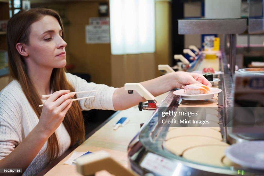 Young Caucasian woman sitting in a sushi bar, choosing a dish from a sushi train, Kaiten-zushi.