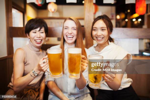 three women sitting side by side at a table in a restaurant, holding large glasses with beer. - 女子会　日本 ストックフォトと画像