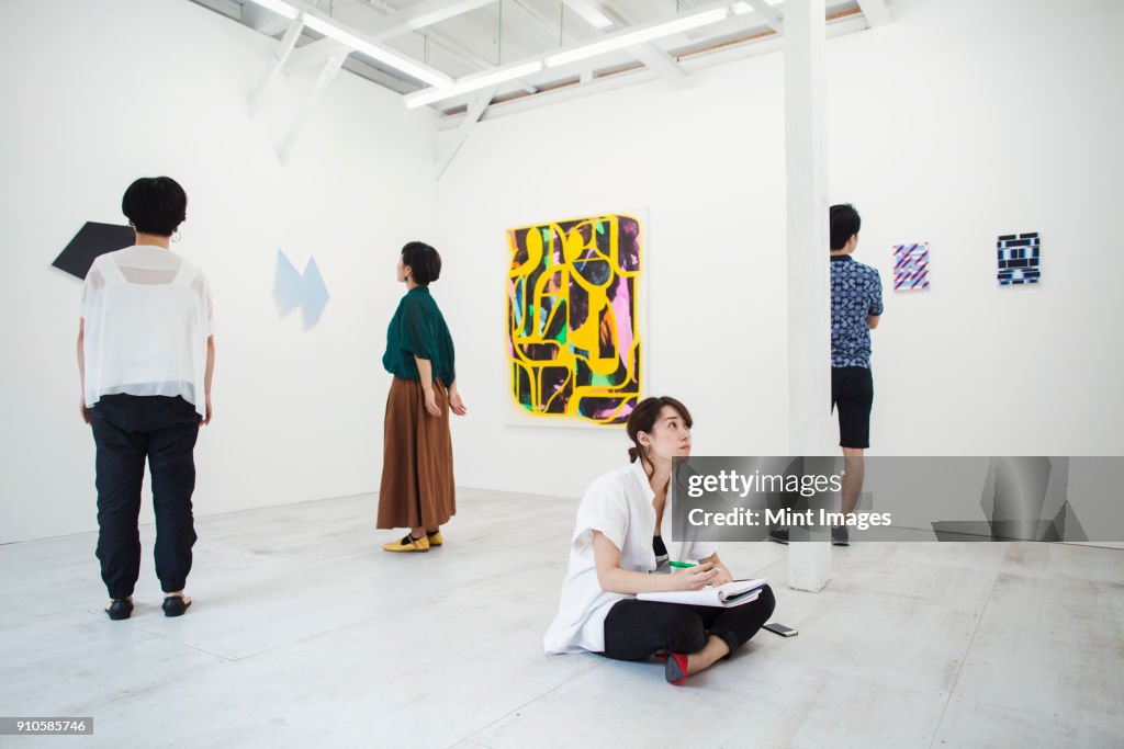Woman with black hair sitting on floor in art gallery with pen and paper, looking at modern painting, three people standing in front of artworks.