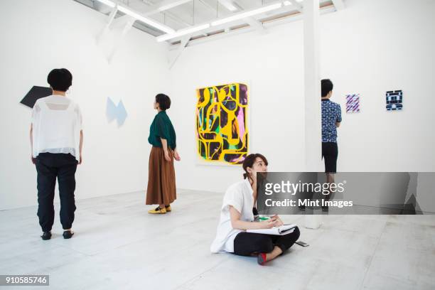 woman with black hair sitting on floor in art gallery with pen and paper, looking at modern painting, three people standing in front of artworks. - modern art stock pictures, royalty-free photos & images