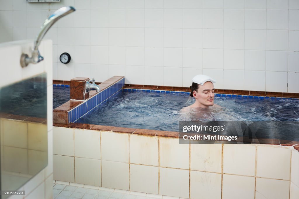 A man in a public bath house bath, in deep water with moving water jets, eyes closed.