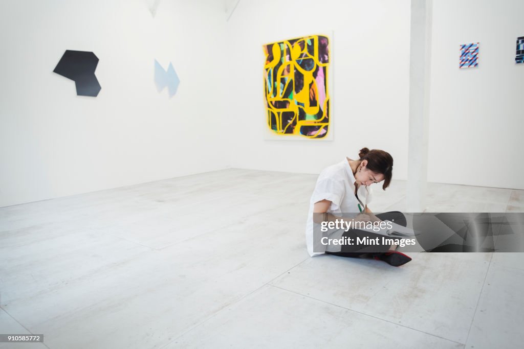 Woman with black hair wearing white shirt sitting on floor in art gallery with pen and paper.