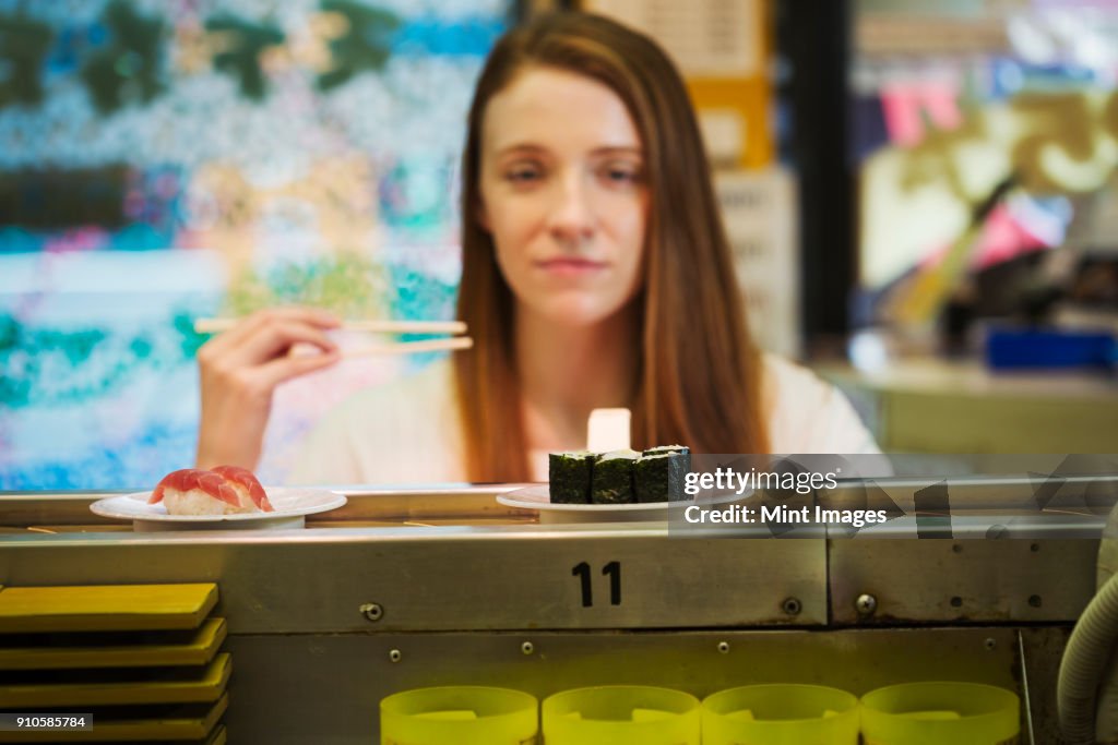 Young woman sitting in a sushi bar, choosing a dish from a sushi train, Kaiten-zushi.
