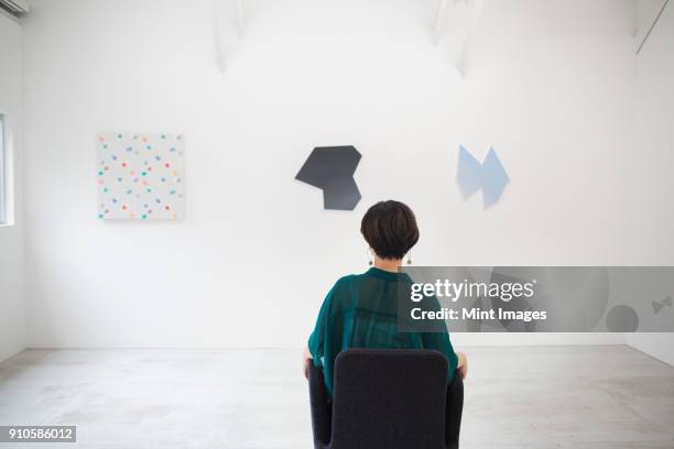 rear view of woman with short black hair wearing green shirt sitting in art gallery, looking at modern paintings. - asian woman short hair stock pictures, royalty-free photos & images