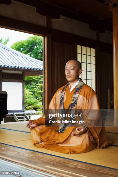 buddhist monk with shaved head wearing golden robe sitting cross legged on the floor, meditating, buddhist hand gesture. - monk stock-fotos und bilder