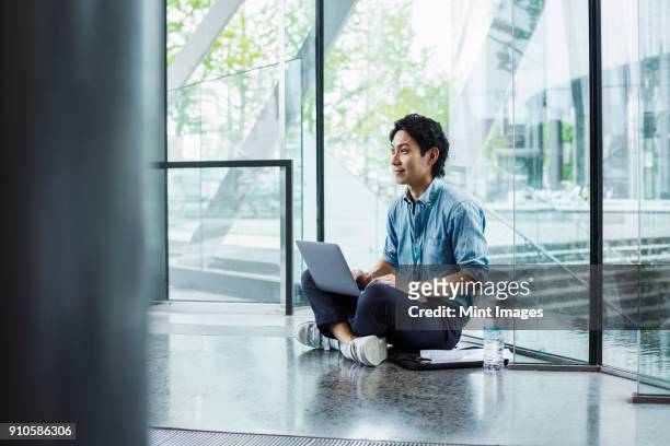 businessman wearing blue shirt sitting on floor indoors, leaning against glass wall, working on laptop computer. - sitting on ground stock pictures, royalty-free photos & images