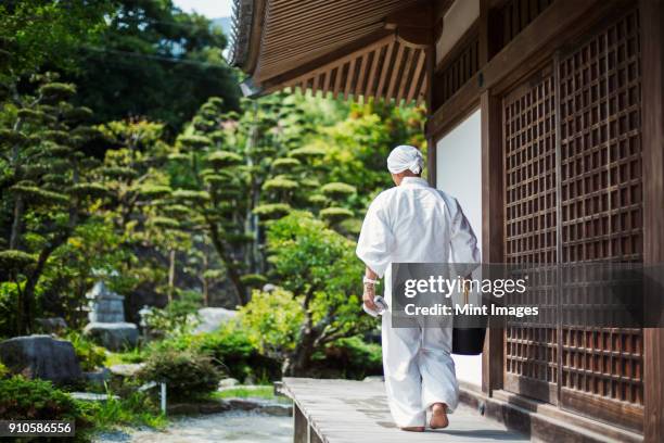 rear view of buddhist monk wearing white robe and cap walking outside a temple, carrying black plastic bucket. - shingon buddhismus stock-fotos und bilder