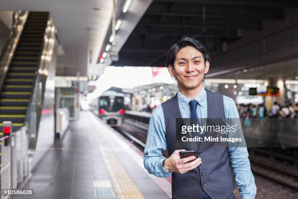 businessman wearing blue shirt and vest standing on train station platform, holding mobile phone, looking at camera. - station ストックフォトと画像
