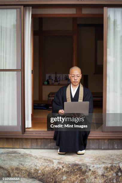 buddhist monk with shaved head wearing black robe sitting outside temple, looking at laptop computer. - shingon buddhismus stock-fotos und bilder