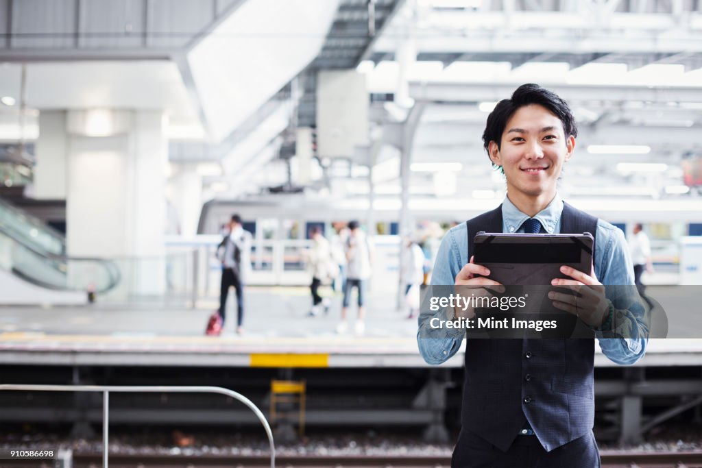 Businessman wearing blue shirt and vest standing on train station platform, holding digital tablet, looking at camera.