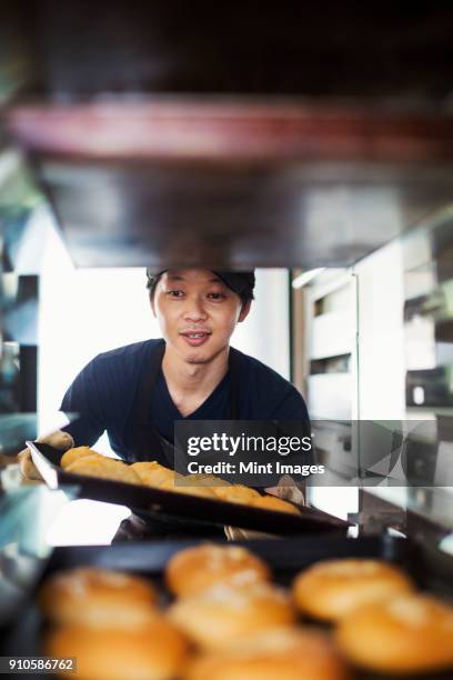 man working in a bakery, placing large trays with freshly baked rolls on a trolley. - man tray food holding stock pictures, royalty-free photos & images