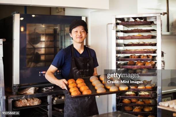 man working in a bakery, holding large tray with freshly baked rolls, smiling at camera. - food and drink establishment stock pictures, royalty-free photos & images