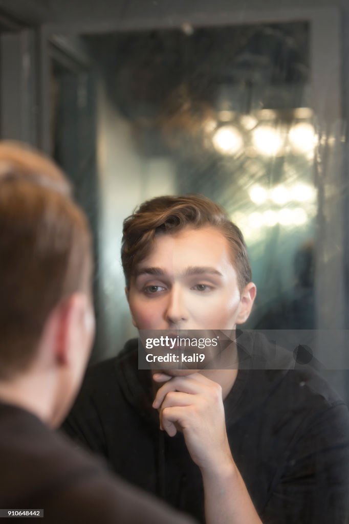 Young man in dressing room staring at reflection, wearing make-up