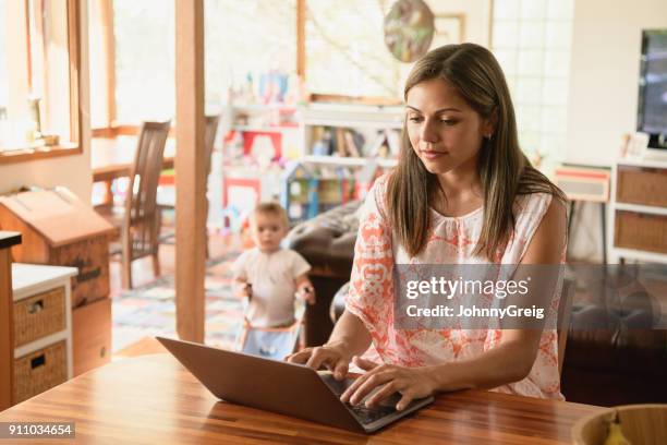 young woman using laptop with baby son in background - adult baby women stock pictures, royalty-free photos & images