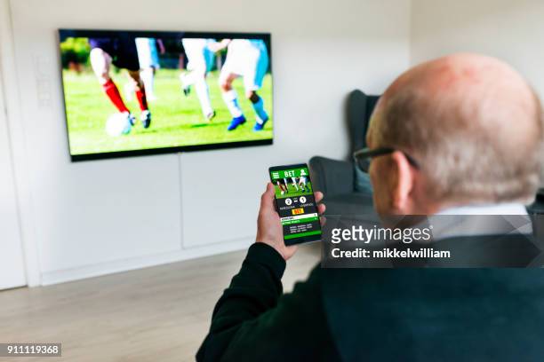 man watches soccer match on television and bets on the game with betting app on phone - betting football sport stock pictures, royalty-free photos & images