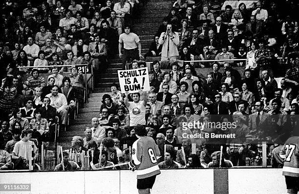 Dave Schultz of the Philadelphia Flyers skates in front of his bench as a fan of the New York Islanders holds a sign that reads "Schultz is a bum"...