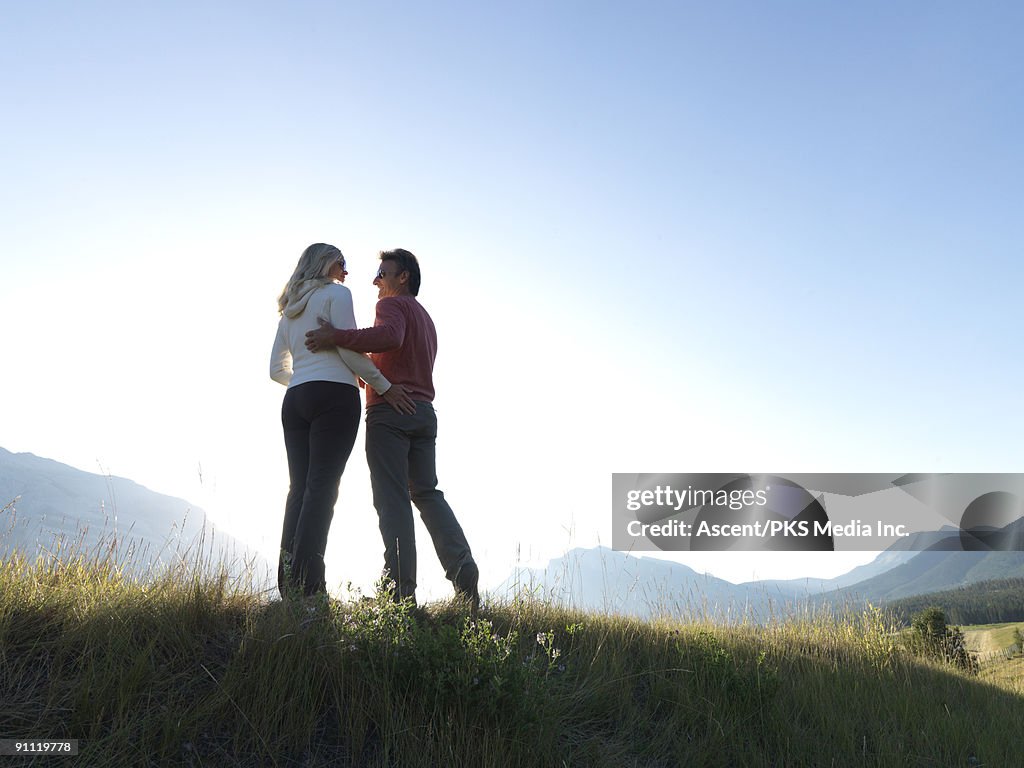 Couple stand on grassy ridge crest, arms holding