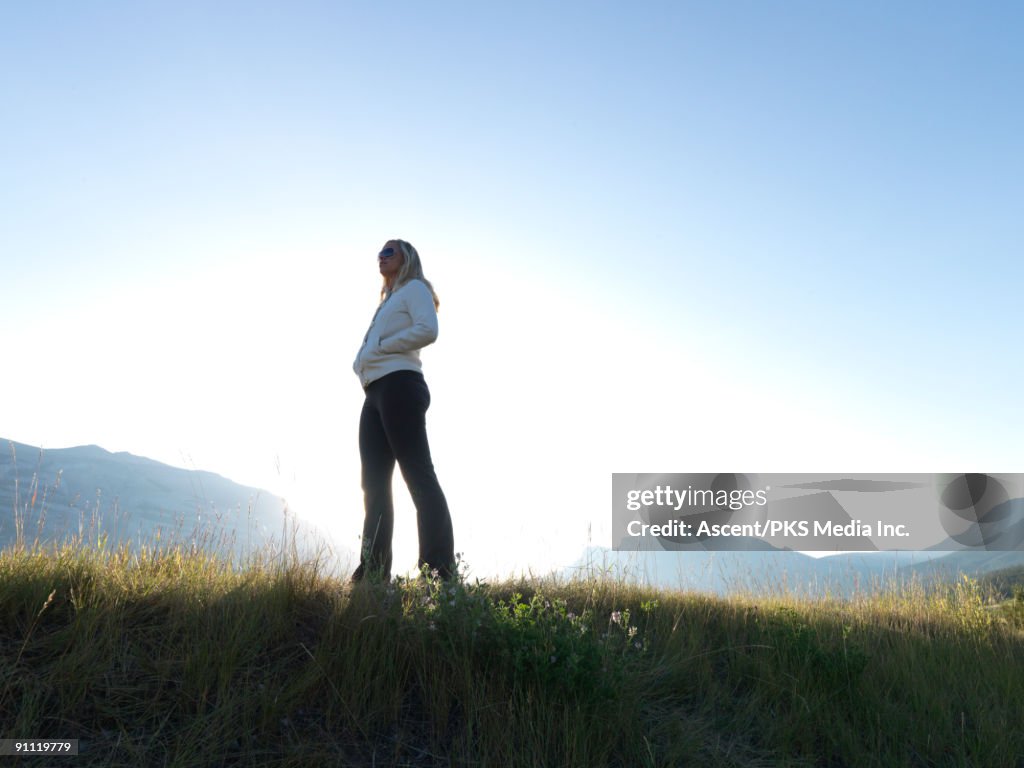 Woman stands on grassy crest, looks towards mtns