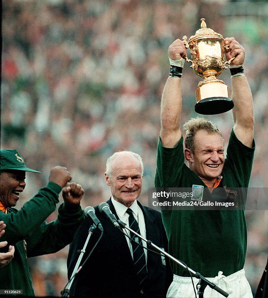 South Africa. Captain of the Springbok team, Francois Pienaar holds the Web Ellis trophy up high after defeating the All Blacks at the World Cup. President Nelson Mandela dances in joy.