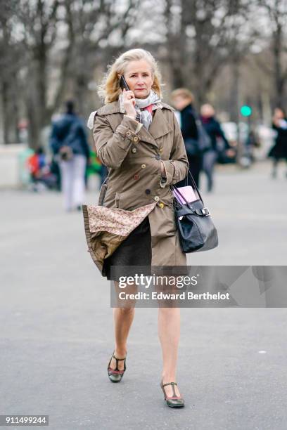 Guest wears a trench coat, a black leather bag, sandals, a scarf, is using a smartphone, outside Chanel, during Paris Fashion Week -Haute Couture...