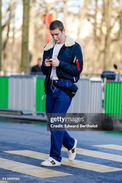 Guest wears a bomber jacket with sheep wool, blue pants, Gucci sneakers, a white t-shirt, is sending a text message with a smartphone, outside...