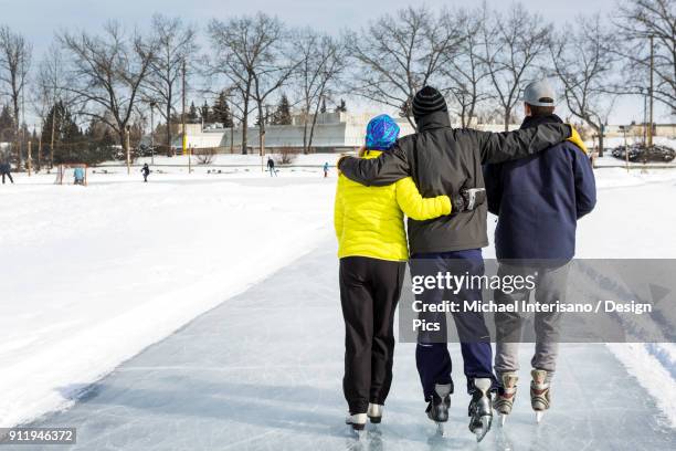 two males and one female skating arm in arm on freshly groomed ice on pond with community centre in the background - family skating in pond foto e immagini stock