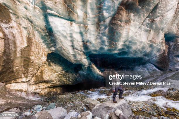 a man surrounded by rushing water poses in front of a cave beneath the ice of root glacier in wrangell-st. elias national park - root glacier stock-fotos und bilder