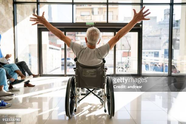 paziente anziano irriconoscibile che lascia la clinica su una sedia a rotelle con le braccia alzate - partire foto e immagini stock