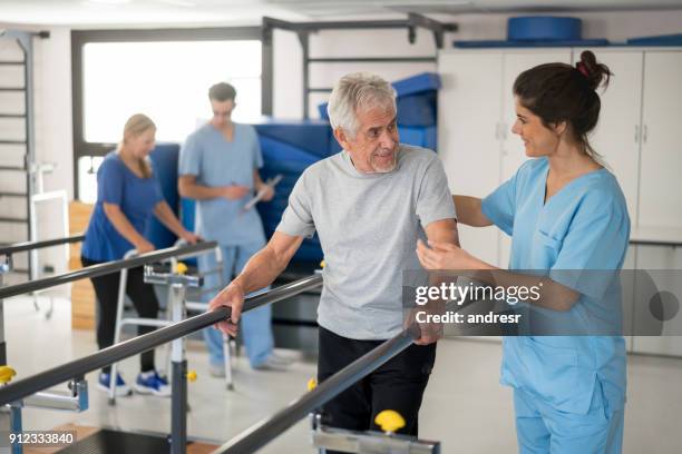 cheerful senior man using parallel bars to walk and physiotherapist smiling at him happy - recuperação imagens e fotografias de stock